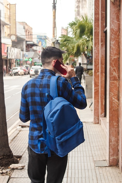 Vista trasera de un hombre vestido con una mochila y una camisa a cuadros azul caminando por las calles de la ciudad mientras realiza una llamada telefónica. - foto vertical.