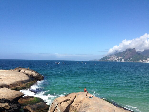 Foto vista trasera de hombres relajándose en una roca en la playa contra el cielo azul