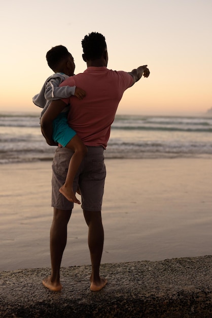 Vista trasera del joven afroamericano que lleva a su hijo y apunta hacia el mar contra el cielo despejado