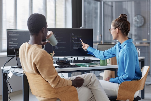 Vista trasera de una joven apuntando al monitor y discutiendo códigos informáticos con colegas