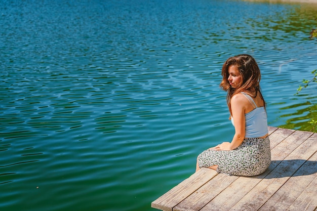 Vista trasera Una joven y bella mujer está sentada en un muelle de madera en un encantador lago de montaña azul