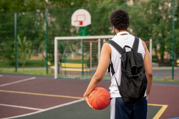 Vista trasera del joven deportista con mochila negra y pelota llegando a la cancha de baloncesto para jugar