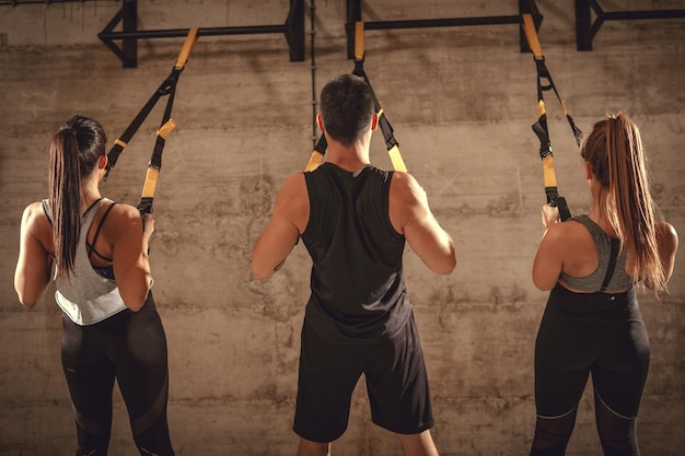 Vista trasera de un joven musculoso y dos mujeres haciendo ejercicio con cinturones en el gimnasio.