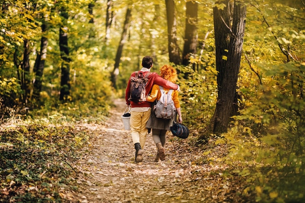 Vista trasera de la joven pareja feliz en el amor abrazándose y caminando en la naturaleza en un hermoso día de otoño. Pareja sosteniendo equipo de picnic.