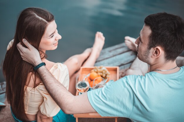 Vista trasera de la joven pareja de picnic cerca de un río o lago, una mujer y un hombre bebiendo vino juntos al aire libre, gente divirtiéndose en las vacaciones de verano