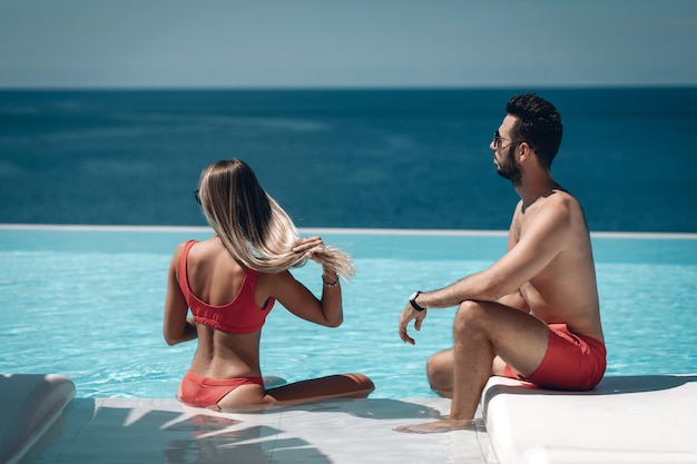 Vista trasera de la joven pareja en traje de baño rojo sentado junto a la piscina en un día soleado. Vacaciones. Phuket. Tailandia