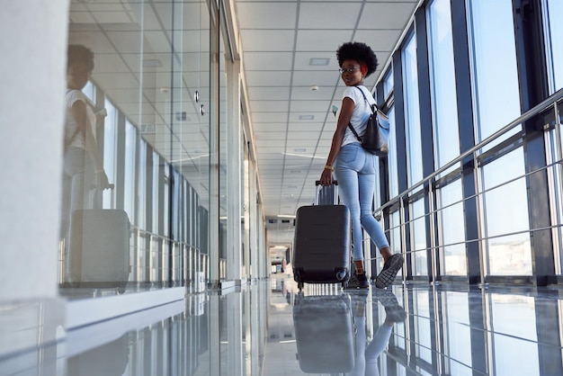 Vista trasera de una joven pasajera afroamericana con ropa informal que está en el aeropuerto con equipaje.