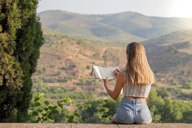 Vista trasera de una joven rubia irreconocible dibujando en un cuaderno el hermoso paisaje