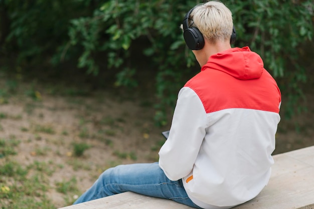 Foto vista trasera del joven rubio caucásico sentado en la pared y escuchando música al aire libre en el parque concepto de estilo de vida