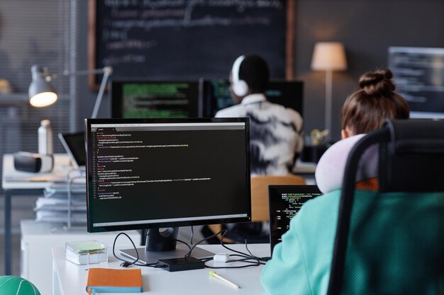 Vista trasera de jóvenes sentados en sus lugares de trabajo con computadoras y trabajando con programas