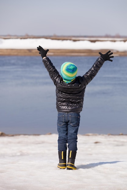 Vista trasera de un lindo niño feliz parado en la orilla del río con las manos en alto en el soleado día de invierno