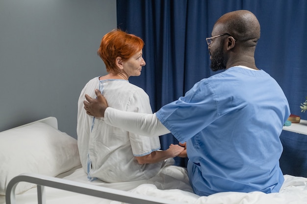 Vista trasera del médico varón africano en uniforme abrazando y hablando con una mujer madura mientras están sentados en la cama en la sala