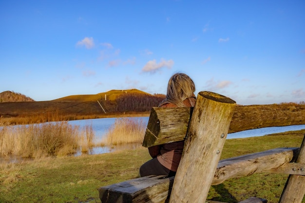 Foto vista trasera de una mujer adulta sentada en un banco de madera con un paisaje otoñal en el fondo del lago y las montañas contra el cielo azul parque thor parque nacional hoge kempen día soleado en genk bélgica