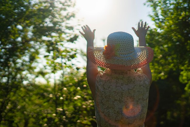 Vista trasera de una mujer adulta con sombrero de verano levantando la mano para que brille el sol