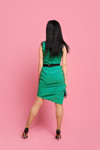 Vista trasera de una mujer de cabello oscuro con vestido verde sentado sobre fondo rosa