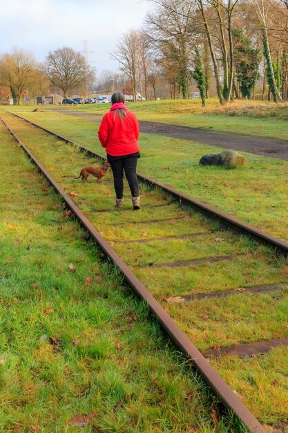 Foto vista trasera de una mujer caminando con su perro entre los rieles de las vías del tren en desuso de la antigua estación as, traviesas cubiertas de hierba, autos estacionados y árboles desnudos en el fondo, soleado día de otoño en limburgo, bélgica