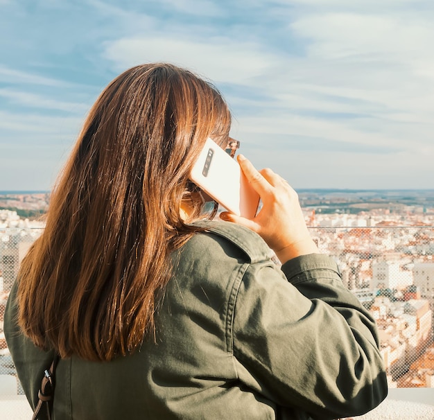 Vista trasera de una mujer caucásica hablando por su smartphone desde la azotea de un edificio.