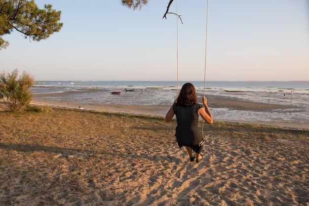 Vista trasera de una mujer en un columpio en una playa mirando el océano