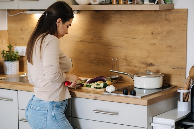 Vista trasera de una mujer cortando diferentes verduras para preparar un plato vegano en su cocina