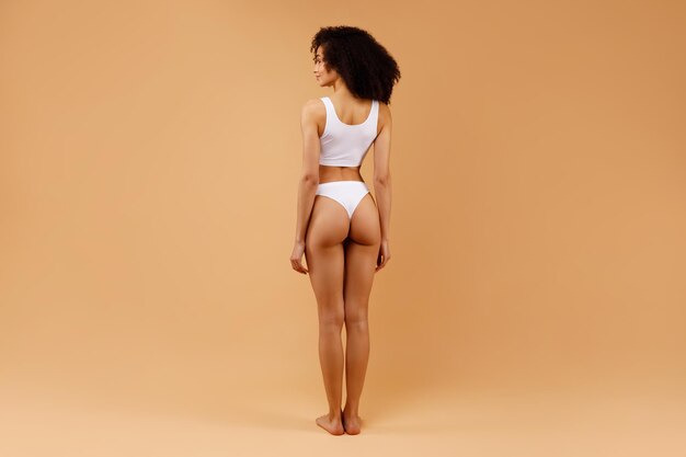 Foto vista trasera de una mujer esbelta en ropa interior blanca que demuestra su cuerpo perfecto fondo beige completo