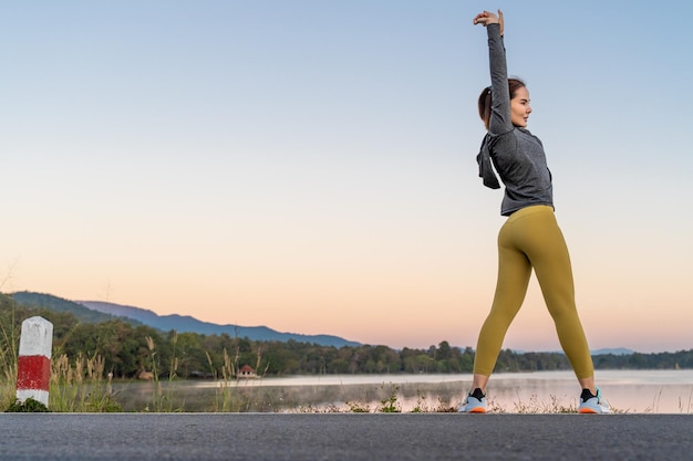 Foto vista trasera de una mujer estirando sus brazos y piernas antes de su ejercicio matutino en un parque de un lago local