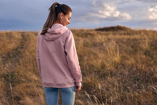 Vista trasera de la mujer feliz en sudadera con capucha rosa y leggins de pie en el campo por la mañana. vista desde atrás en la dama de pelo largo que va a entrenar al aire libre al aire libre, al amanecer. concepto de estilo de vida de la gente