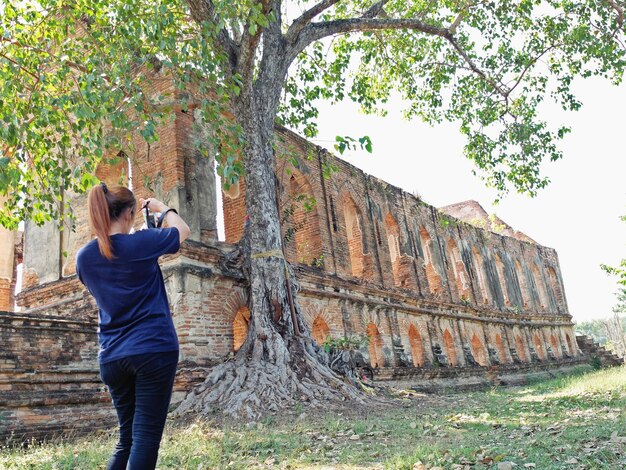 Foto vista trasera de una mujer fotografiando un árbol junto a antiguas ruinas