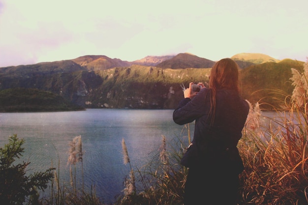 Vista trasera de una mujer fotografiando el lago y las montañas a través del teléfono
