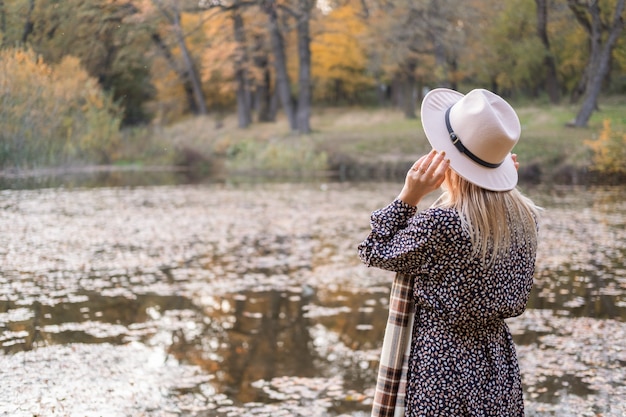 Vista trasera de una mujer hermosa en ropa de moda y sombrero junto al lago en el parque de otoño en otoño.