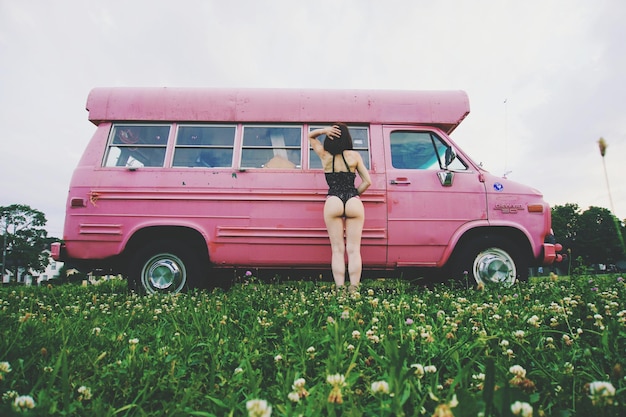 Vista trasera de una mujer joven en bikini de pie junto al coche en un campo de hierba contra un cielo despejado