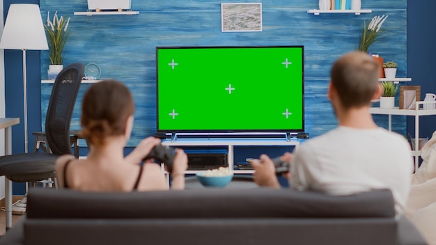 Vista trasera de una mujer joven y un novio que sostienen controladores jugando un juego de acción en la consola en la televisión de pantalla verde sentados en el sofá. Pareja pasando tiempo libre jugando en la pantalla de maqueta de clave cromática.