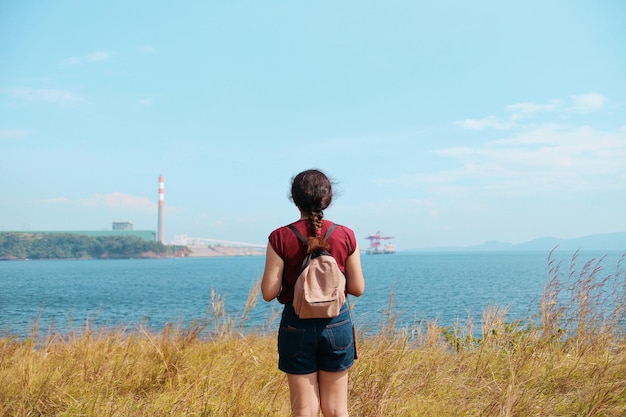Vista trasera de una mujer joven de pie en el campo por el mar contra el cielo