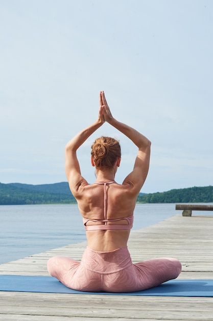 Vista trasera mujer madura en ropa deportiva rosa practica yoga en una plataforma de madera cerca del mar