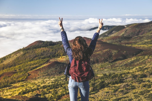 Vista trasera de una mujer con mochilero gestando un signo de paz mientras mira el paisaje