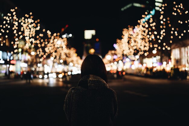 Foto vista trasera de una mujer de pie en una calle de la ciudad contra las luces de navidad por la noche