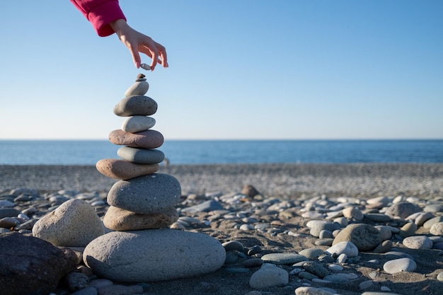 Vista trasera de una mujer de pie en la playa contra un cielo despejado y construyendo la torre de piedras y rocas