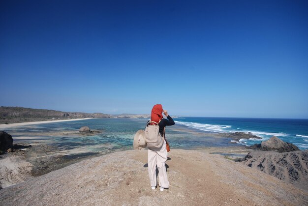 Vista trasera de una mujer de pie en una roca en la playa contra un cielo azul claro