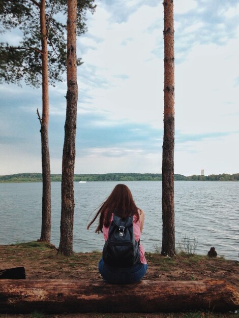 Foto vista trasera de una mujer sentada en un árbol caído en la orilla del río contra el cielo