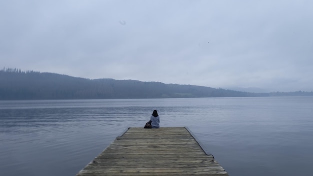 Vista trasera de una mujer sentada en el muelle del lago contra el cielo