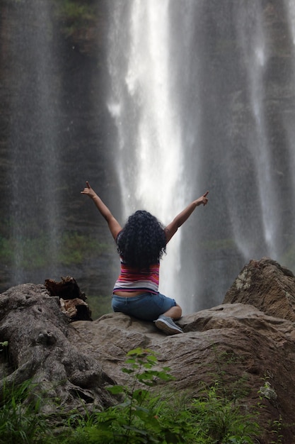 Foto vista trasera de una mujer sentada en una roca contra una cascada