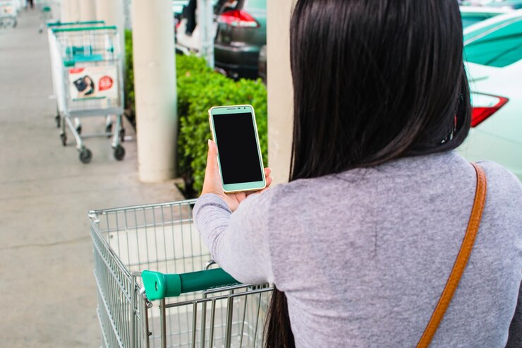 Foto vista trasera de una mujer con un teléfono inteligente junto al carrito de compras al aire libre