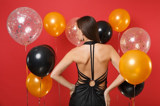 Vista trasera de la mujer en vestido negro celebrando, de pie con los brazos en jarras en globos de aire de fondo rojo brillante. Día internacional de la mujer, feliz año nuevo, concepto de fiesta de vacaciones de maqueta de cumpleaños.