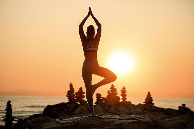 Vista trasera de una mujer zen en pose de árbol meditando en una roca junto al mar al atardecer. Copie el espacio.