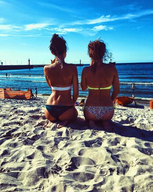 Foto vista trasera de mujeres jóvenes sentadas en la arena en la playa contra el cielo