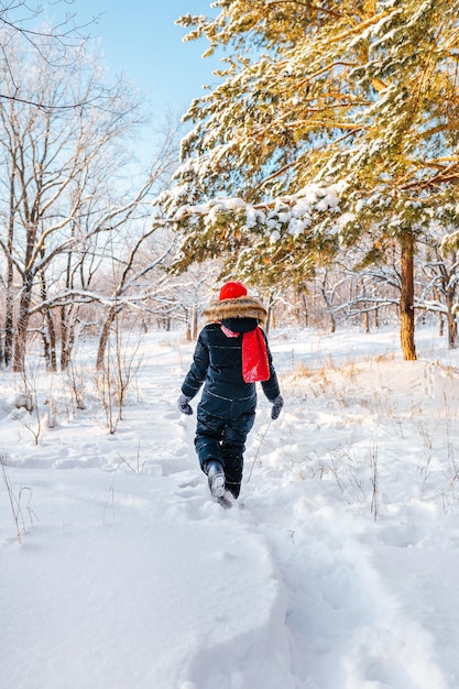Vista trasera de una niña de 10 años con un sombrero rojo en un bosque de invierno con luz solar