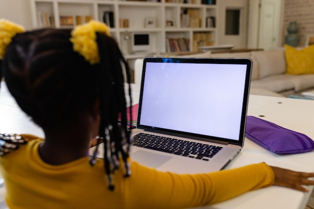 Vista trasera de una niña afroamericana estudiando en línea a través de una computadora portátil en casa, espacio de copia