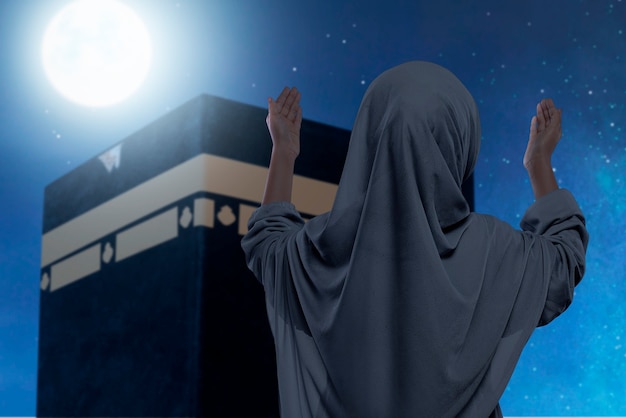 Vista trasera de la niña musulmana asiática en un velo de pie y manos levantadas rezando frente a la Kaaba con el fondo de la escena nocturna