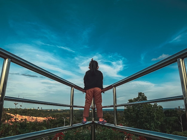 Foto vista trasera de la niña de pie en la barandilla contra el cielo