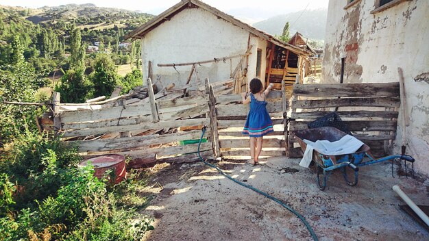 Vista trasera de la niña de pie junto a la valla de madera contra la casa