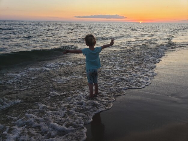 Vista trasera de una niña de pie en la playa durante la puesta de sol